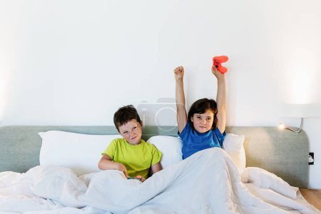 Foto de Chica levantando brazos con la almohadilla de juego en la emoción mientras juega videojuego con el hermano molesto en la cama en el dormitorio de luz - Imagen libre de derechos