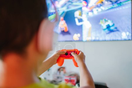 Photo for Back view of cropped anonymous boy in wireless headphones with gamepad playing interesting video game on TV in light living room - Royalty Free Image