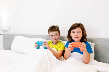 Téléchargez les photos : Frères et sœurs focalisés avec des manettes de jeu jouant jeu vidéo intéressant tout en étant assis ensemble dans un lit confortable sous couverture dans la chambre à coucher à la maison - en image libre de droit