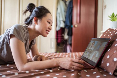 Photo for Side view of young Asian female lying on bed and chatting via video call on laptop with group of friends while spending time at home during coronavirus pandemic - Royalty Free Image
