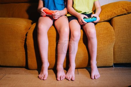 Téléchargez les photos : Frères et sœurs méconnaissables recadrés avec des coussinets de jeu jouant au jeu vidéo assis sur le canapé dans le salon avec une lumière tamisée - en image libre de droit