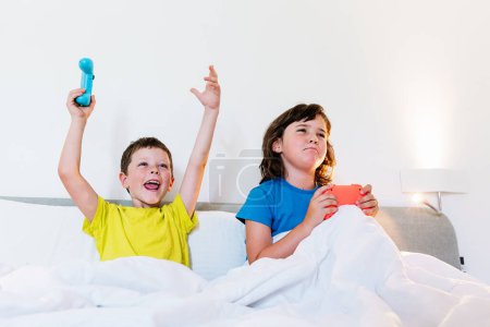 Téléchargez les photos : Garçon levant les bras avec tampon de jeu dans l'excitation tout en jouant au jeu vidéo avec sœur bouleversée au lit dans la chambre à coucher légère - en image libre de droit