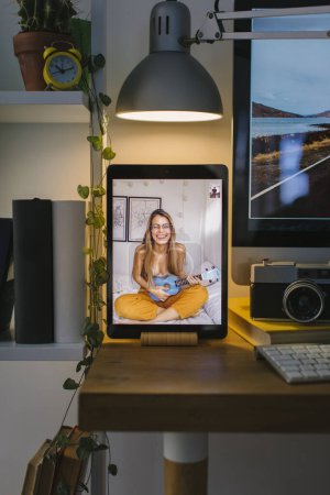 Photo for Happy smiley blonde girl playing ukulele during a video call with a friend in Corona Times. The tablet is in a front view over a desk with a computer, some books and a camera. - Royalty Free Image