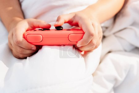 Cropped unrecognizable child with game pad playing video game in bed in light bedroom