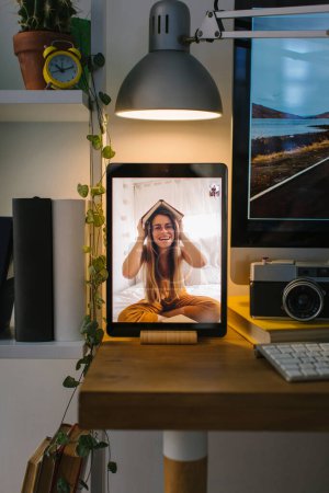 Téléchargez les photos : Adolescente blonde jouant avec un livre lors d'un appel vidéo dans sa chambre. La tablette est dans une vue de face sur un bureau avec un ordinateur, quelques livres et un appareil photo. - en image libre de droit