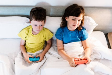 Téléchargez les photos : De ci-dessus frères et sœurs concentrés avec des manettes de jeu jouant jeu vidéo intéressant tout en étant assis ensemble dans un lit confortable sous couverture dans la chambre à coucher à la maison - en image libre de droit