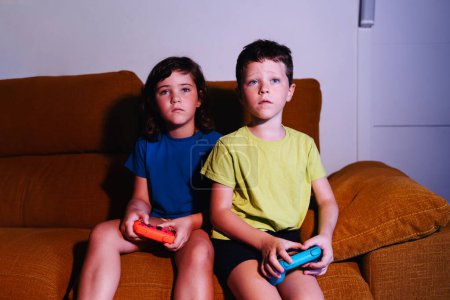Photo for Focused siblings with game pads playing video game while sitting on couch in living room with dim light looking away - Royalty Free Image