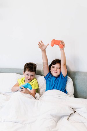 Téléchargez les photos : Contenu bras levant avec tapis de jeu dans l'excitation tout en jouant avec frère intéressant jeu vidéo tout en étant assis ensemble dans un lit confortable sous couverture dans la chambre à coucher à la maison - en image libre de droit