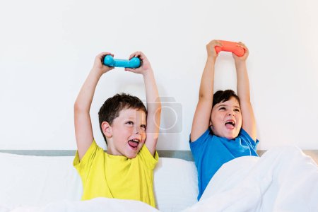Photo for Content siblings raising arms with game pad in excitement while playing interesting video game while sitting together in comfortable bed under blanket in bedroom at home - Royalty Free Image