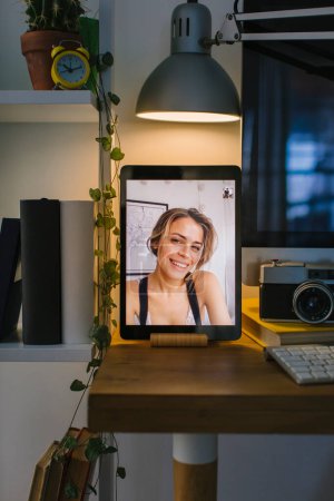 Girl smiling to her friend during a video call in Corona Times. The tablet is in a front view over a desk with a computer, some books and a camera.