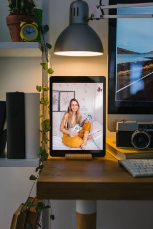 Téléchargez les photos : Joyeux fille jouant un ukulélé bleu dans son lit lors d'un appel vidéo. La tablette est dans une vue de face sur un bureau avec un ordinateur, quelques livres et un appareil photo. - en image libre de droit