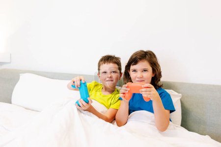 Téléchargez les photos : Contenu frères et sœurs avec des manettes de jeu jouant jeu vidéo intéressant tout en étant assis ensemble dans un lit confortable sous couverture dans la chambre à coucher à la maison - en image libre de droit