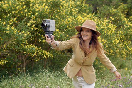 Téléchargez les photos : Femme mince souriante en tenue décontractée et chapeau flânant avec une vieille caméra vidéo près des buissons avec des fleurs en fleurs colorées à la campagne et regardant loin - en image libre de droit