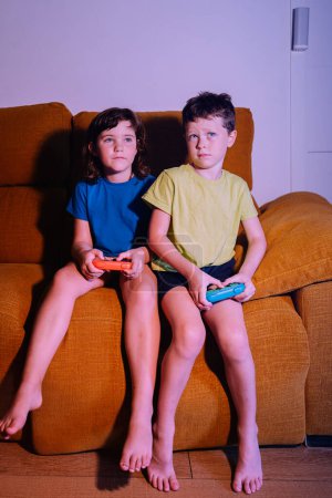 Foto de Cuerpo completo de hermanos enfocados con almohadillas de juego jugando videojuego mientras están sentados en el sofá en la sala de estar con poca luz - Imagen libre de derechos