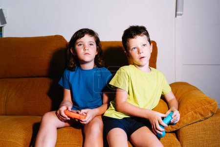 Foto de Hermanos enfocados con almohadillas de juego jugando videojuego mientras están sentados en el sofá en la sala de estar con poca luz mirando hacia otro lado - Imagen libre de derechos