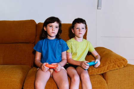 Téléchargez les photos : Frères et sœurs focalisés avec des coussinets de jeu jouant au jeu vidéo assis sur le canapé dans le salon avec une lumière tamisée regardant loin - en image libre de droit