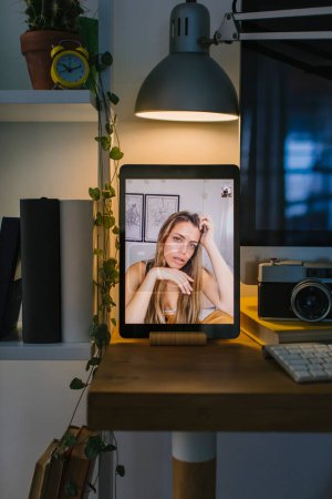 Portrait of a blonde teenager during a video call with a photographer in Coronavirus quarantine. The tablet is in a front view over a desk with a computer, some books and a camera.