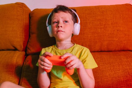 Photo for Focused boy in wireless headphones with gamepad while playing video game on couch in living room looking away - Royalty Free Image