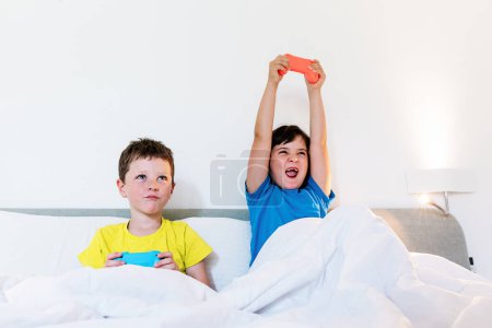 Foto de Chica levantando brazos con la almohadilla de juego en la emoción mientras juega videojuego con el hermano molesto en la cama en el dormitorio de luz - Imagen libre de derechos