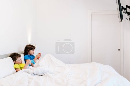 Foto de Vista lateral de los hermanos enfocados con gamepads jugando interesante videojuego mientras están sentados juntos en una cómoda cama bajo una manta en el dormitorio en casa - Imagen libre de derechos