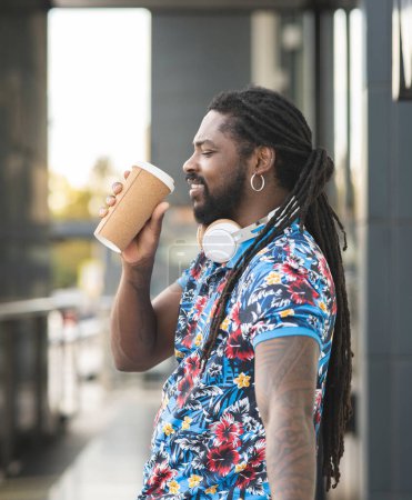 Téléchargez les photos : Vue latérale d'un homme afro-américain tatoué avec des dreadlocks debout dans la rue avec un casque sans fil sur le cou et une boisson chaude à emporter - en image libre de droit