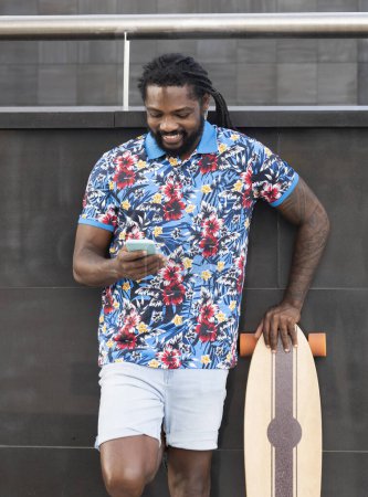 Foto de Alegre barbudo hombre afroamericano en ropa casual surfeando teléfono celular mientras está de pie cerca de la pared del edificio con longboard en la calle de la ciudad - Imagen libre de derechos