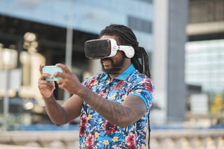Photo for African American male with dreadlocks in contemporary VR goggles surfing cellphone while exploring virtual reality on street with modern buildings - Royalty Free Image