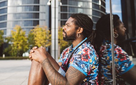 Photo for Side view of serious thoughtful bearded African American male in dreadlocks and casual clothes looking at camera while sitting near modern building with reflection in city - Royalty Free Image
