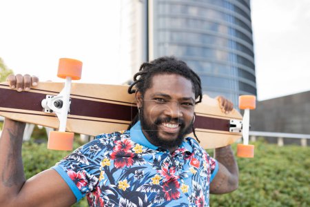 Photo for Delighted African American male in casual clothes with longboard behind head looking at camera while standing on street of city - Royalty Free Image