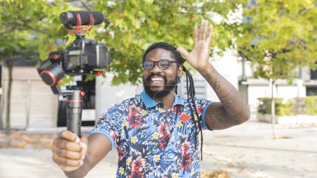 Photo for African American blogger with dreadlocks in eyeglasses waving hands while shooting video on professional camera on street with trees in city - Royalty Free Image