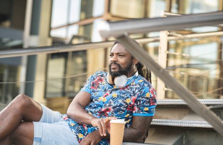 Téléchargez les photos : Homme afro-américain réfléchi en vêtements décontractés avec casque sans fil sur le cou regardant au loin tout en étant assis sur les escaliers près du bâtiment avec boisson chaude à emporter - en image libre de droit