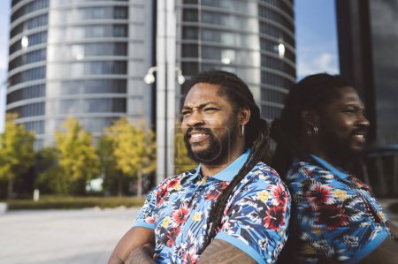 Foto de Feliz barbudo hombre afroamericano en rastas y ropa casual mirando hacia otro lado mientras está sentado cerca de un edificio moderno con reflejo en la ciudad - Imagen libre de derechos