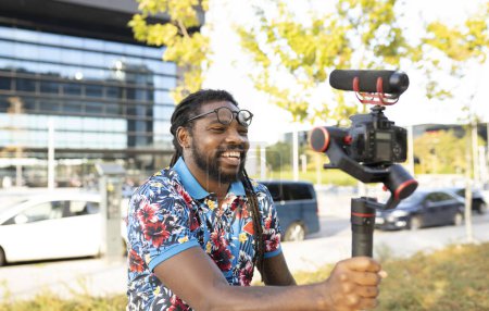 Téléchargez les photos : Blogueur afro-américain avec dreadlocks dans les lunettes tournage vidéo sur caméra professionnelle dans la rue avec des arbres en ville - en image libre de droit