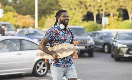 Photo for Cheerful African American male with wireless headphones carrying longboard and takeaway coffee walking near parking lot with modern cars in city - Royalty Free Image