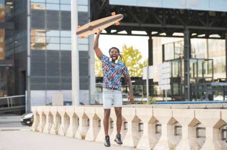 Foto de Emocionado hombre afroamericano con auriculares inalámbricos saltando con longboard en la mano en la pasarela pavimentada en la ciudad - Imagen libre de derechos