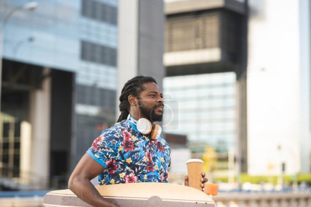 Téléchargez les photos : Vue latérale de l'homme afro-américain avec écouteurs sans fil transportant longboard et café à emporter marchant près du parking avec des voitures modernes en ville - en image libre de droit