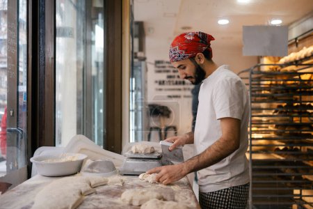 Photo for Young arabic baker dressed with white clothes and a red hat cutting dough with a scapula and weighing it on a scale in a bakery - Royalty Free Image