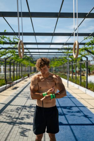 Photo for Handsome young man with blond hair shirtless putting on wrist straps to exercise on the rings outdoors on a sunny day - Royalty Free Image