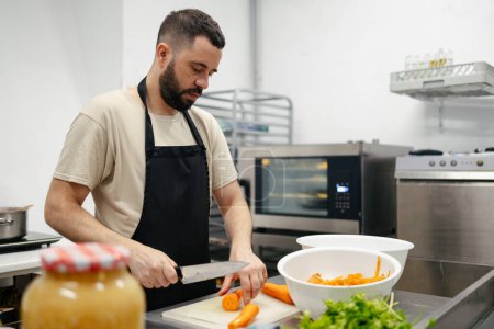 Photo for Young bearded chef dressed in a kitchen apron cutting carrots in the table of a well-equipped kitchen - Royalty Free Image