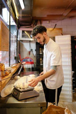Foto de Joven pizzero árabe en delantal blanco que pesa masa en una balanza en la cocina de una pizzería - Imagen libre de derechos