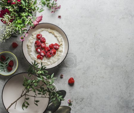 Photo for Romantic breakfast with porridge and heart of raspberries on grey kitchen table with flowers and bowls. Vegan breakfast idea with nutritious ingredients. Top view with copy space. - Royalty Free Image