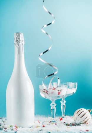 Photo for Christmas and New Year setting with champagne bottle, glasses, falling silver ribbon, snow and bauble and at light blue background. Front view - Royalty Free Image