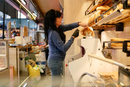 Téléchargez les photos : Femme adulte aux cheveux noirs et aux vêtements bleus mettant une miche de pain dans un sac après l'avoir tranchée pour un client dans une boulangerie - en image libre de droit