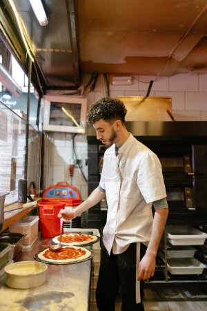 Photo for Young arabic pizza man in white apron adding tomato sauce to the base of a pizza in the kitchen of a pizzeria - Royalty Free Image