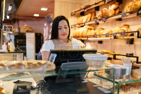 Photo for Young latina woman with black hair and white t-shirt touching the screen of a cash register in a bakery - Royalty Free Image