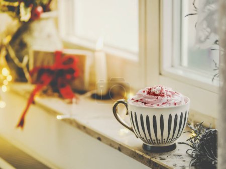 Photo for Mug with hot chocolate and whipped cream on window sill with candle, present and warm light. Hygge winter at home with warming drink. - Royalty Free Image