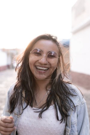 Téléchargez les photos : Une jeune femme hispanique portant des lunettes de soleil sourit et pose dans la rue au coucher du soleil. Concept de photoshoot style de vie en plein air - en image libre de droit