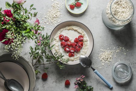Photo for Bowl with porridge and heart shaped raspberries at grey concrete kitchen table with glass jar, branches, bowls and spoon. Preparing healthy vegan breakfast at home. Top view. - Royalty Free Image