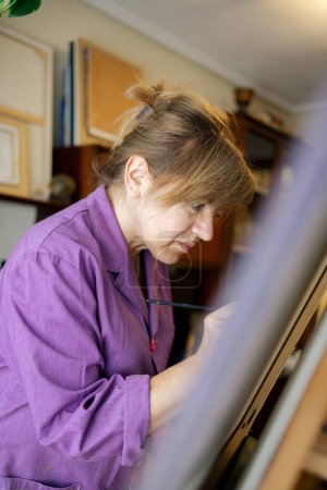 Téléchargez les photos : Femme adulte aux cheveux blonds vêtue d'une robe violette peint au pinceau sur toile dans sa maison - en image libre de droit