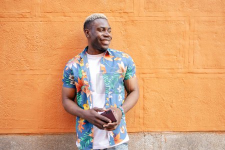 Téléchargez les photos : Portrait d'un beau jeune homme noir portant des vêtements décontractés regardant vers sa gauche en souriant tout en tenant son smartphone appuyé contre un mur orange - en image libre de droit